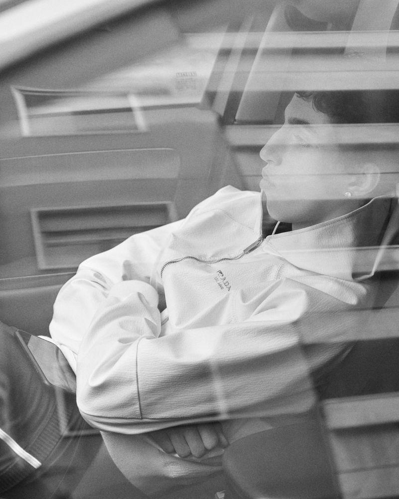 Portrait of a young adult wearing a light hooded jacket, captured through a window with blinds, creating a layered, reflective monochrome image.