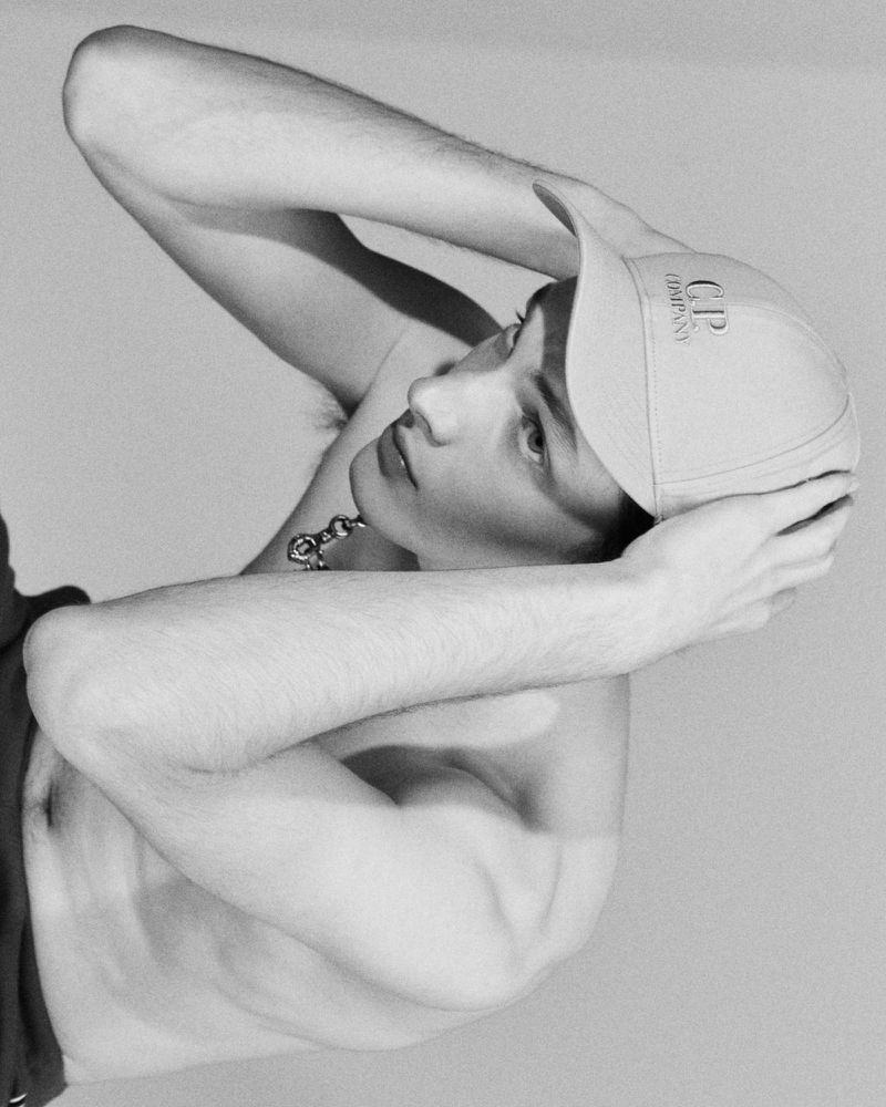 Monochrome portrait of a young adult male wearing a baseball cap, gazing upward with his arms raised above his head.