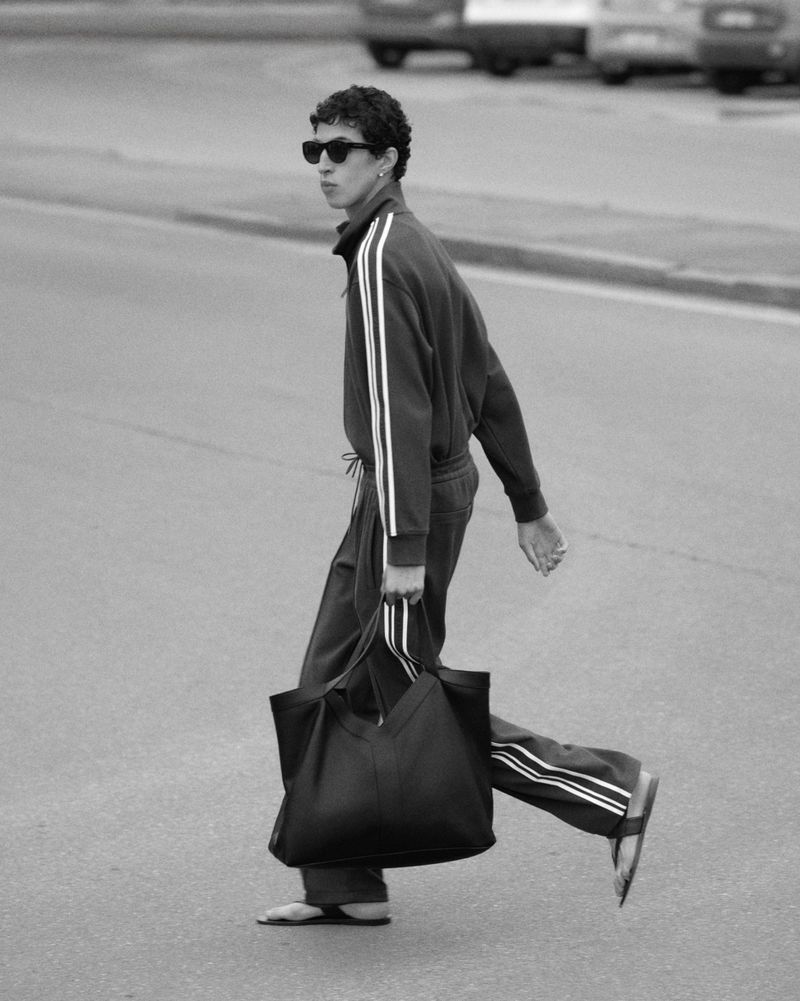 Black-and-white shot of an adult male in a sweatsuit crossing a street while carrying a large tote bag in a monochromatic urban setting.