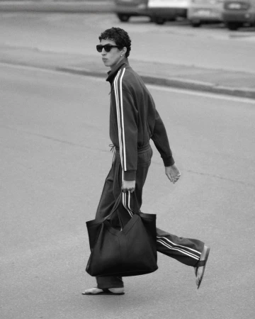 Black-and-white shot of an adult male in a sweatsuit crossing a street while carrying a large tote bag in a monochromatic urban setting.