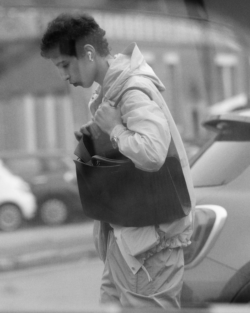 Person with curly hair in a hooded jacket and bag walking on the street near cars in a monochrome, neutral setting.