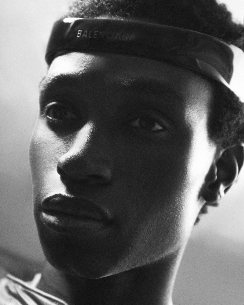 Close-up portrait of a young adult male with short, textured hair wearing a black Balenciaga headband against a softly lit backdrop.