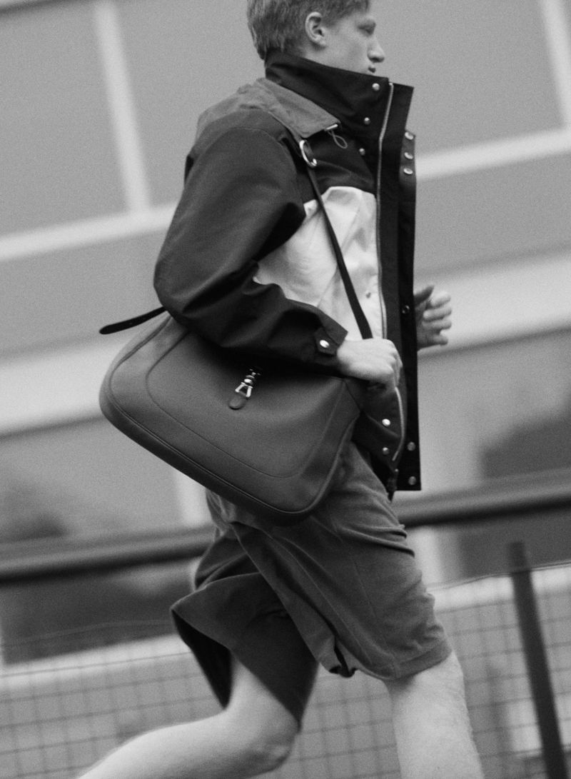 Monochrome shot of a male wearing a jacket and carrying a messenger bag, running outdoors next to a building and metal railing.