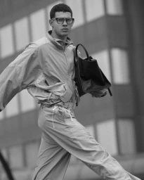 Man wearing glasses and a lightweight tracksuit carrying a dark handbag outdoors