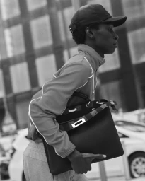 Black model wearing a cap and jacket while carrying a large dark handbag outdoors in a black and white editorial photograph.