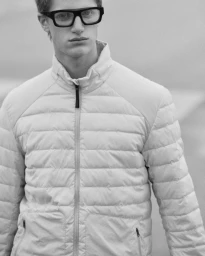 Close-up portrait of a male model wearing black eyeglasses and a light-coloured puffer jacket against a neutral background.