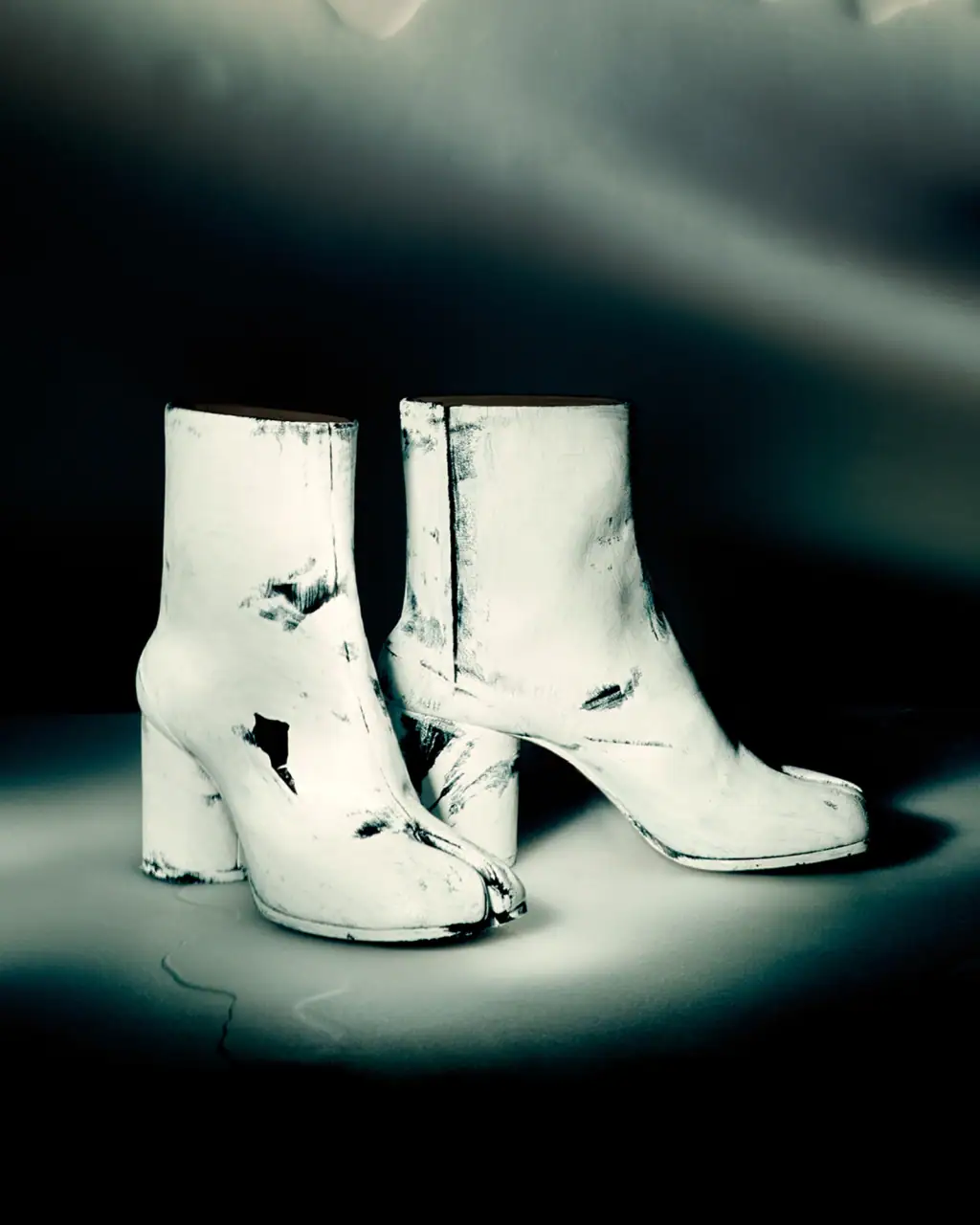Pair of white painted split-toe heeled boots on a dark gradient studio surface for Maison Margiela Fall 2025 campaign.
