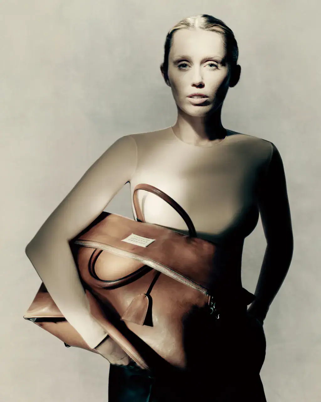 Miley Cyrus holding a large brown leather bag in a Maison Margiela fall ad campaign portrait against a grey background