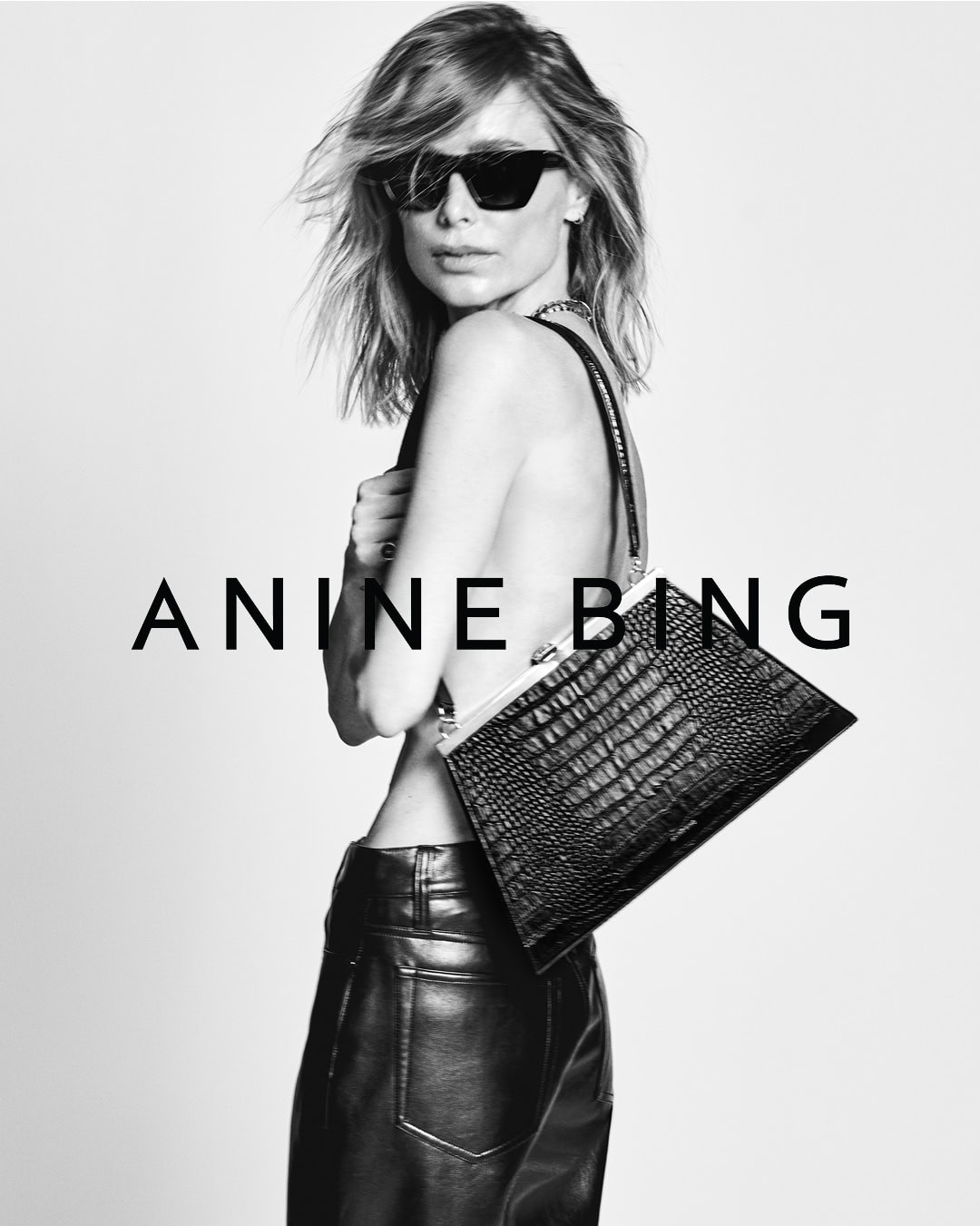 Model in dark sunglasses wearing leather pants and holding a textured handbag against a plain background