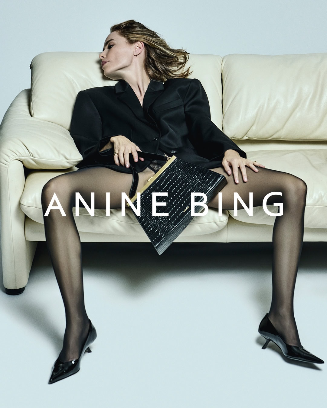 Anine Bing seated on a white leather sofa holding a black embossed handbag for 'The Anine Bag' ad campaign.