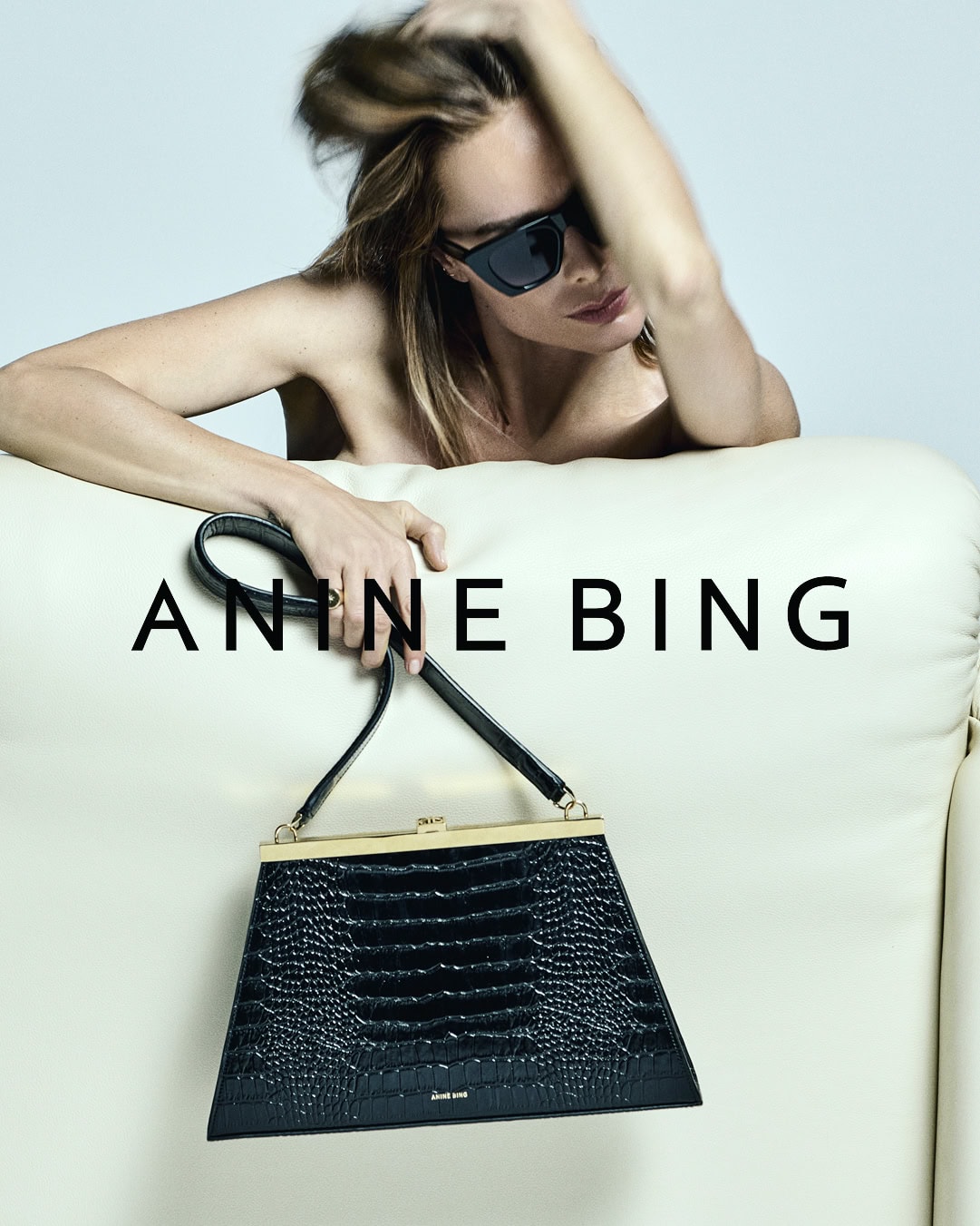 Model wearing dark sunglasses holding a black leather handbag against a light background for Anine Bing
