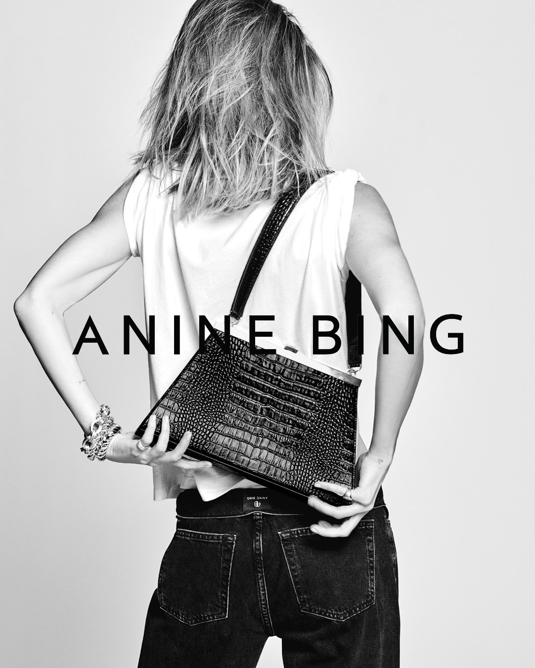 Monochrome fashion campaign image featuring a woman seen from behind wearing a white sleeveless top and dark jeans, holding a black leather handbag.