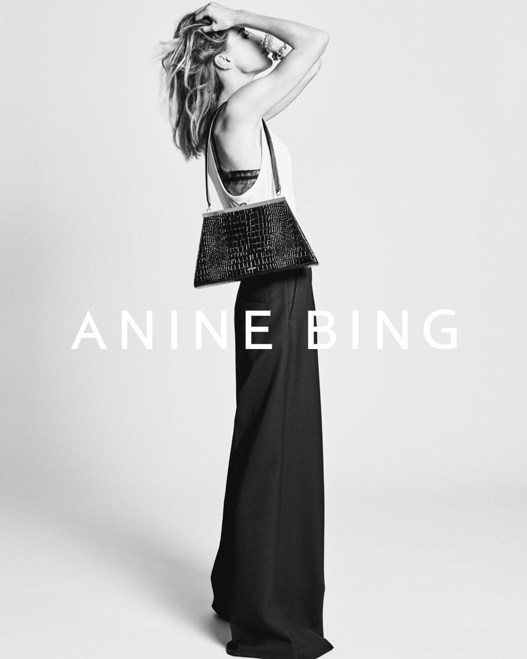 Monochrome fashion campaign image of a model wearing dark trousers holding a textured black handbag against a white seamless backdrop.
