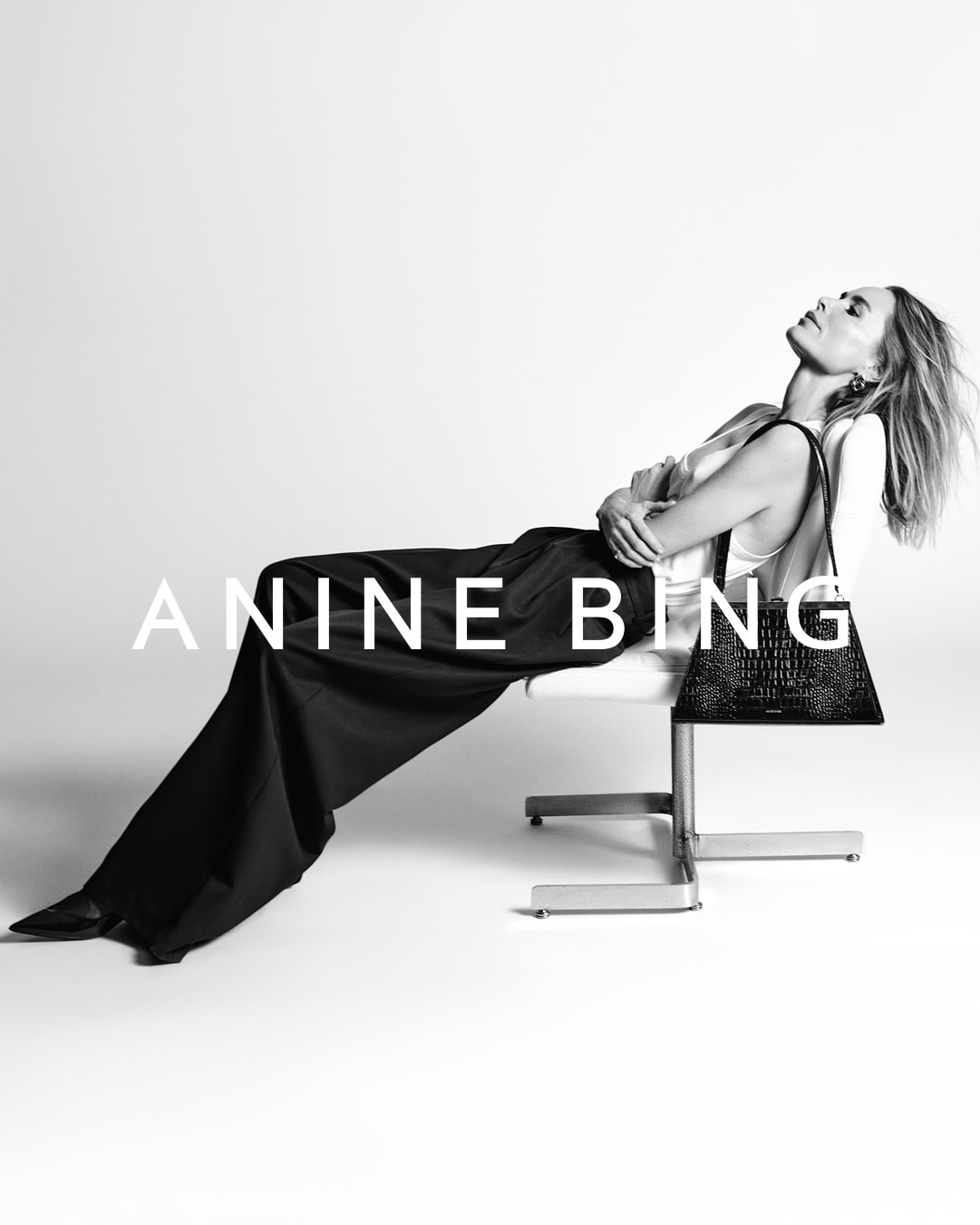 Anine Bing reclining on a white chair in a black trousers and top, holding a black handbag against a seamless background