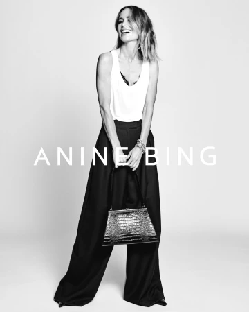 Anine Bing smiling wearing white tank top and black trousers holding a textured handbag against a light seamless background