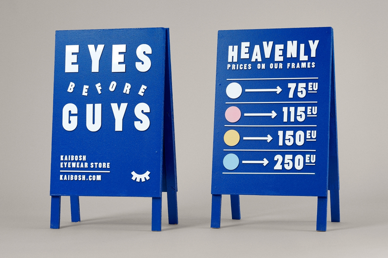 Two blue A-frame wooden signs displaying white lettering and price tiers for Kaibosh eyewear store on a neutral background