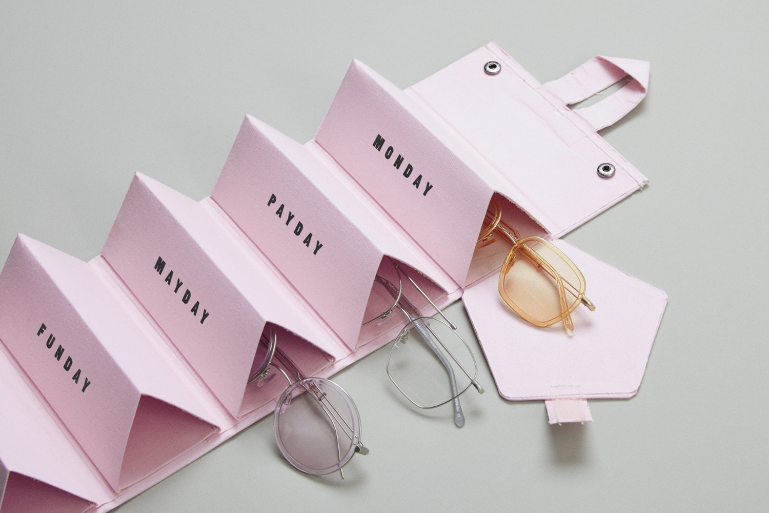 Pink multi-compartment glasses case holding frames, labeled with days, set against a plain grey backdrop.