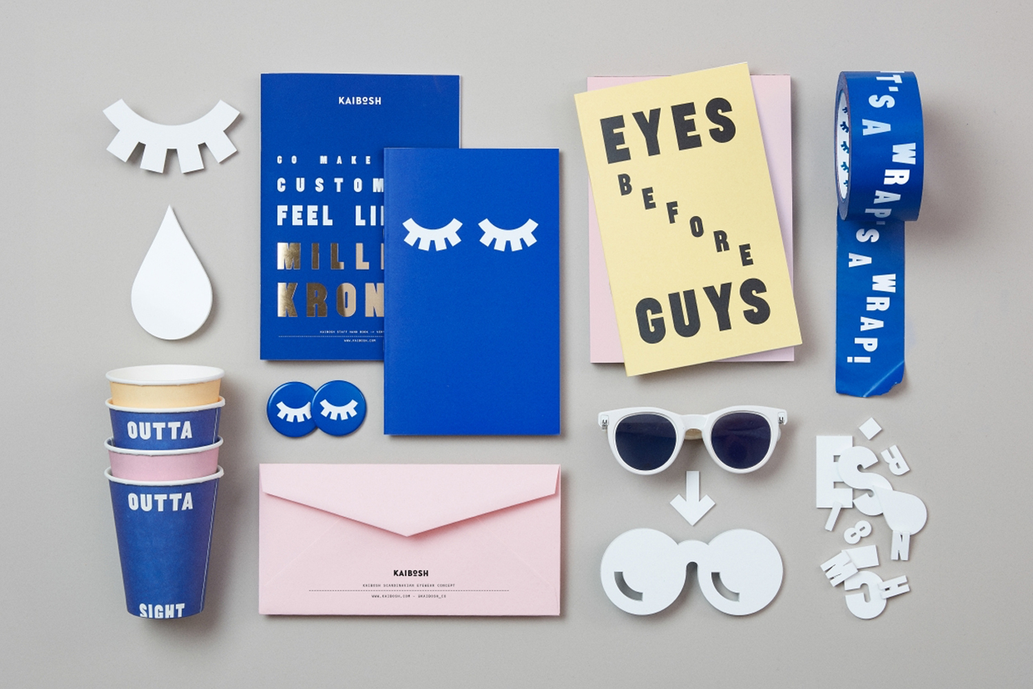Flat lay of Kaibosh brand identity items including blue notebooks, pink envelopes, paper cups, and white sunglasses on a grey backdrop.