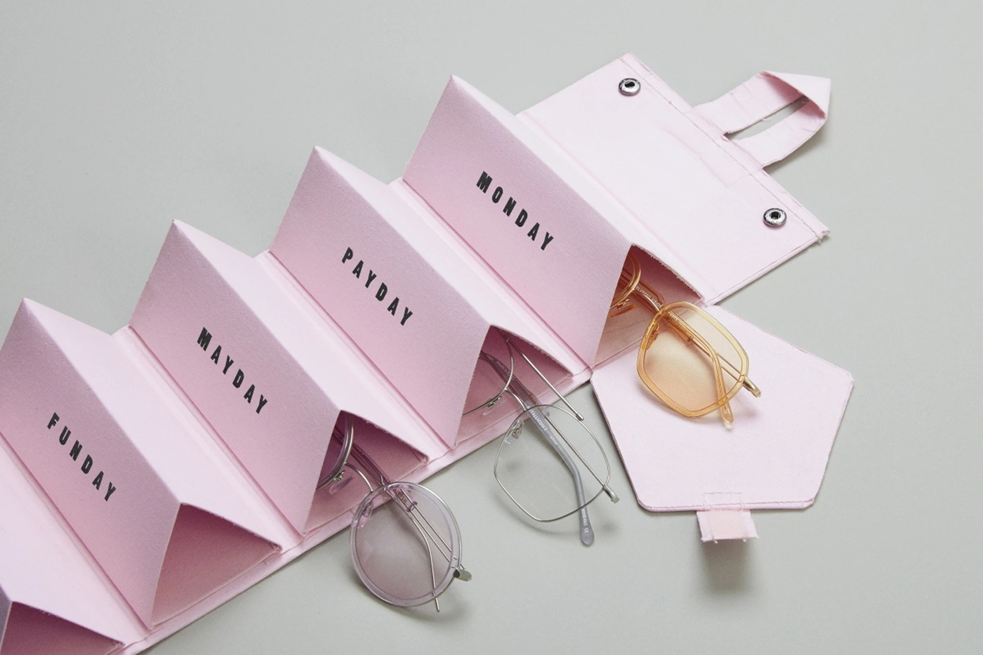 Pink multi-compartment glasses case holding frames, labeled with days, set against a plain grey backdrop.