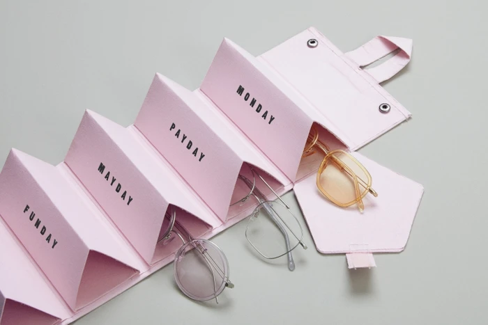 Pink multi-compartment glasses case holding frames, labeled with days, set against a plain grey backdrop.