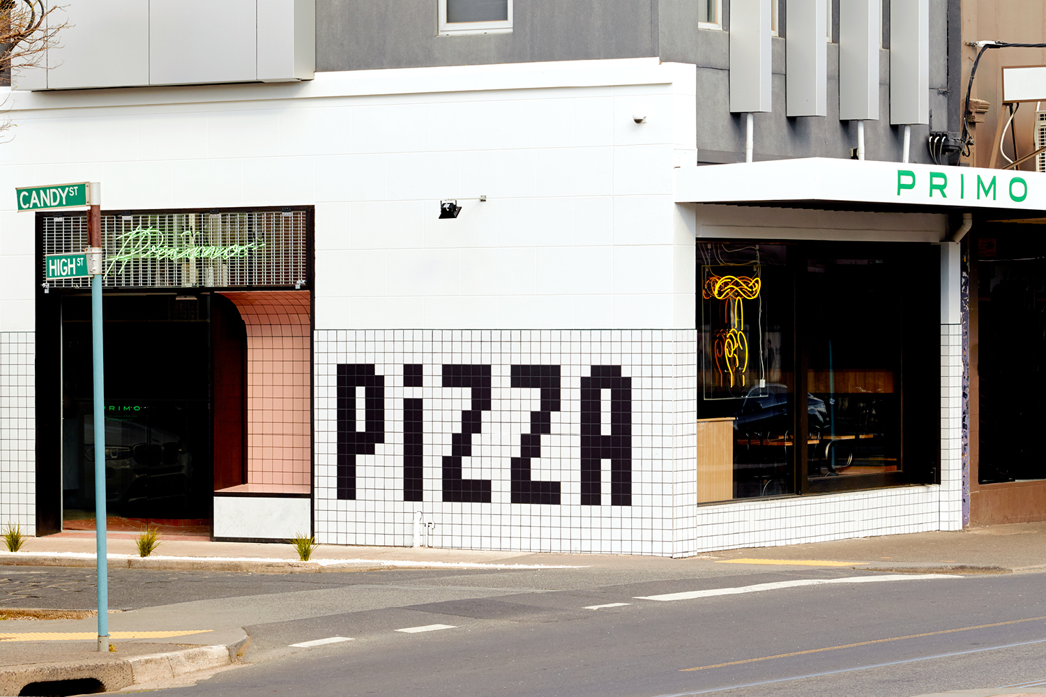 Storefront exterior of Primo featuring black pixelated PIZZA tiles on a white tiled wall, street signs, and large windows