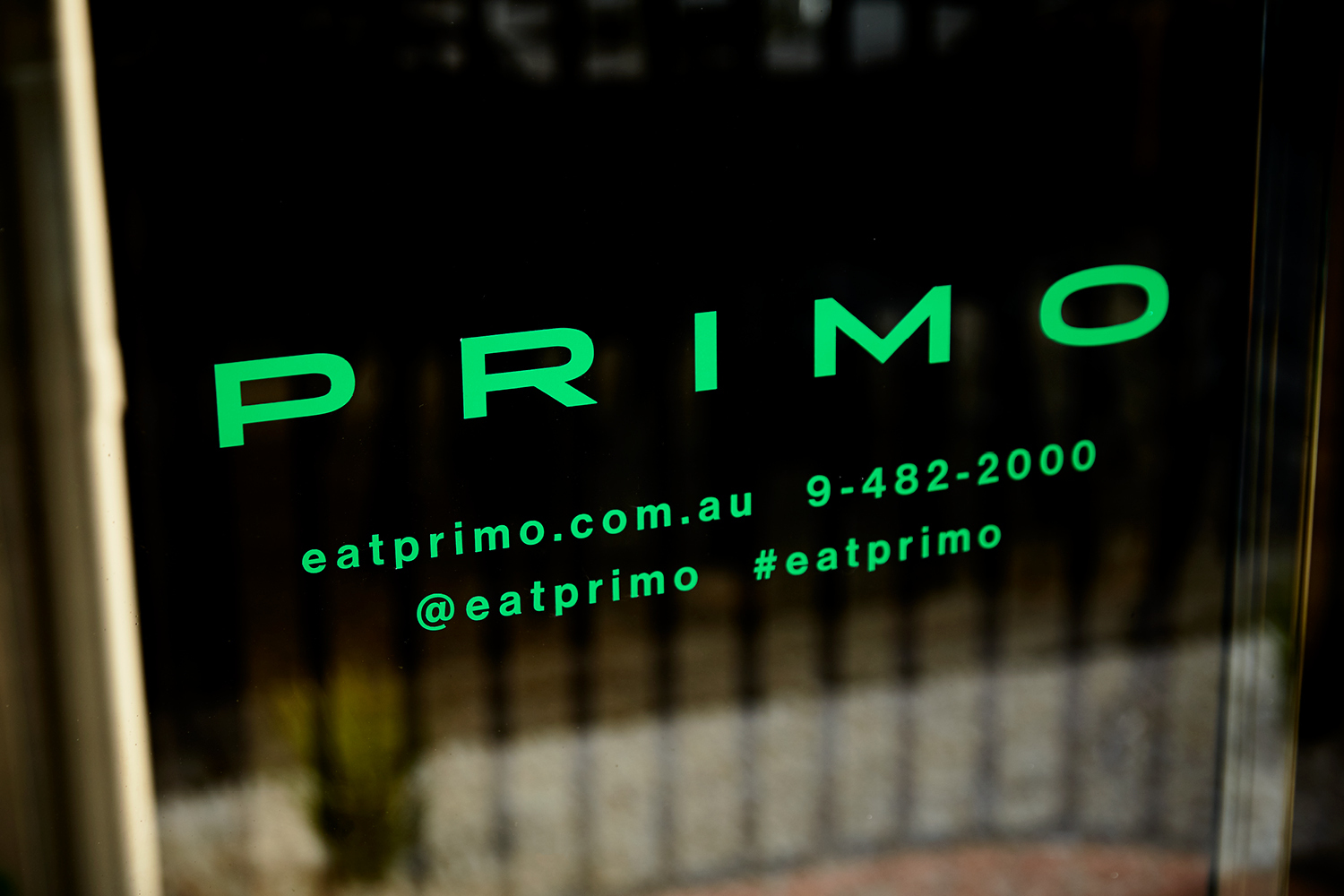 Close-up of bright green Primo brand identity typography printed on dark glass signage with a blurred exterior background.