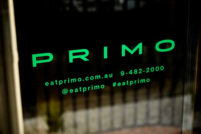 Close-up of bright green Primo brand identity typography printed on dark glass signage with a blurred exterior background.