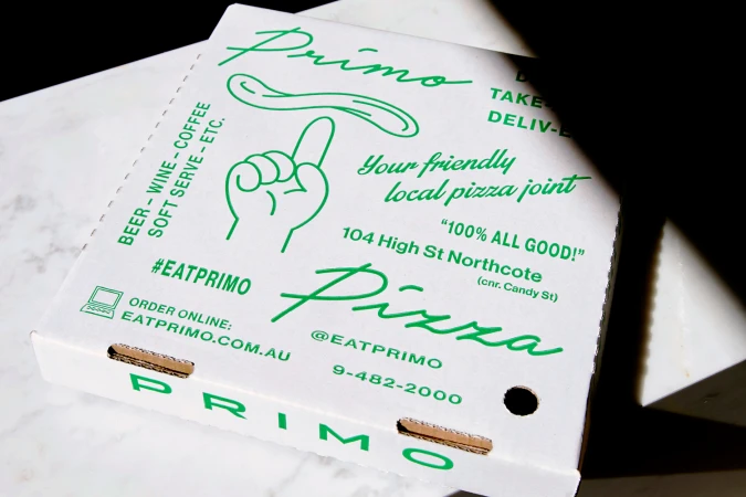 Close-up of a white pizza box with green typography and illustrations, resting on a white marble surface.