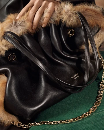 Close-up of a black leather Ferragamo shoulder bag with fur trim, gold chain strap, and hand resting on top.