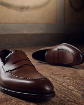 Close-up of brown leather penny loafers resting on a warm wooden floor in a moody indoor setting.