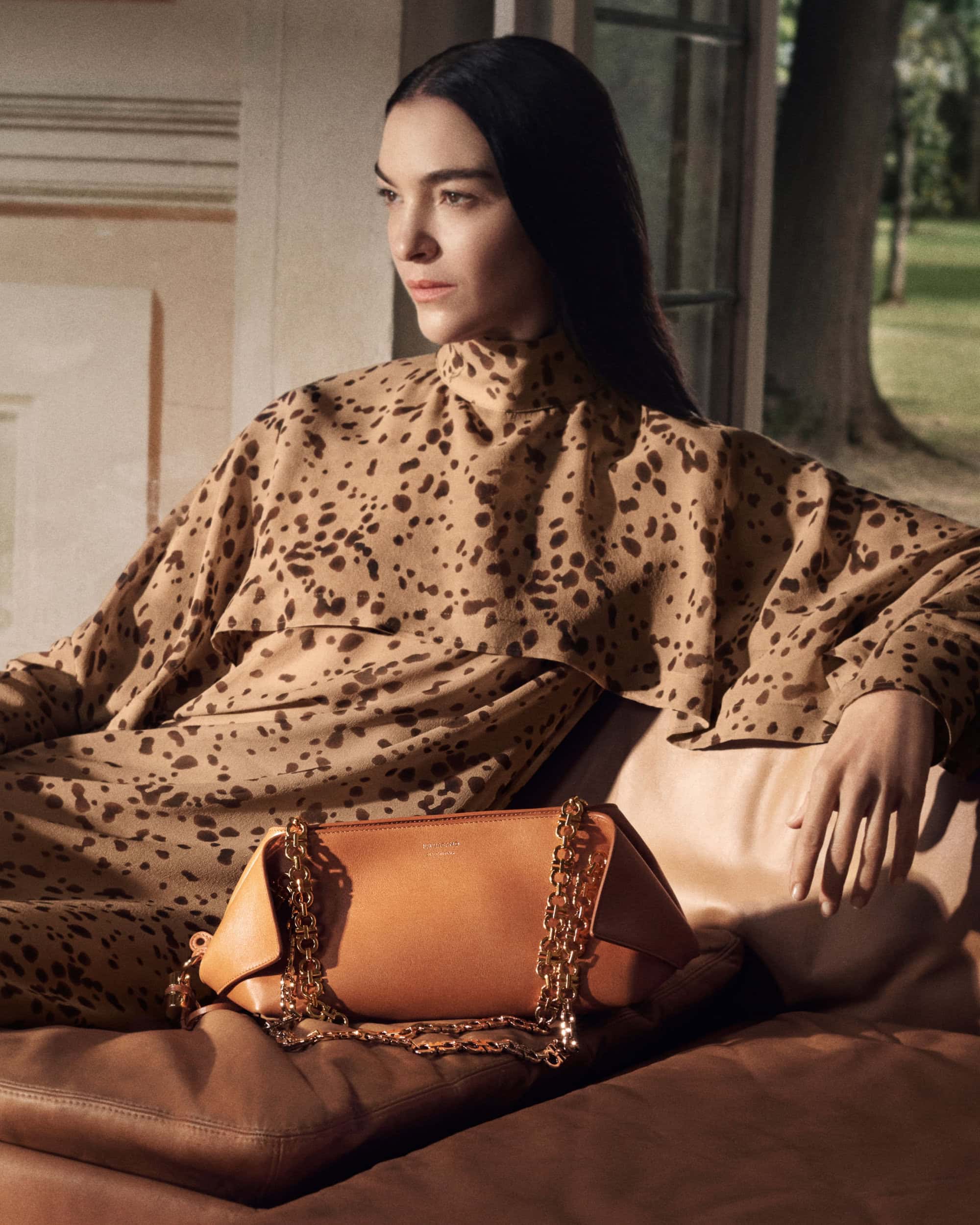 Model seated wearing a printed high-neck garment next to a leather handbag with a chain strap for Ferragamo Fall 2025 campaign.