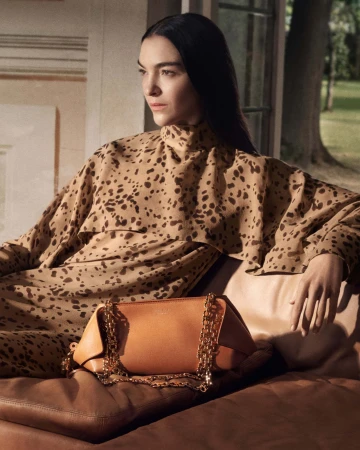 Model seated wearing a printed high-neck garment next to a leather handbag with a chain strap for Ferragamo Fall 2025 campaign.