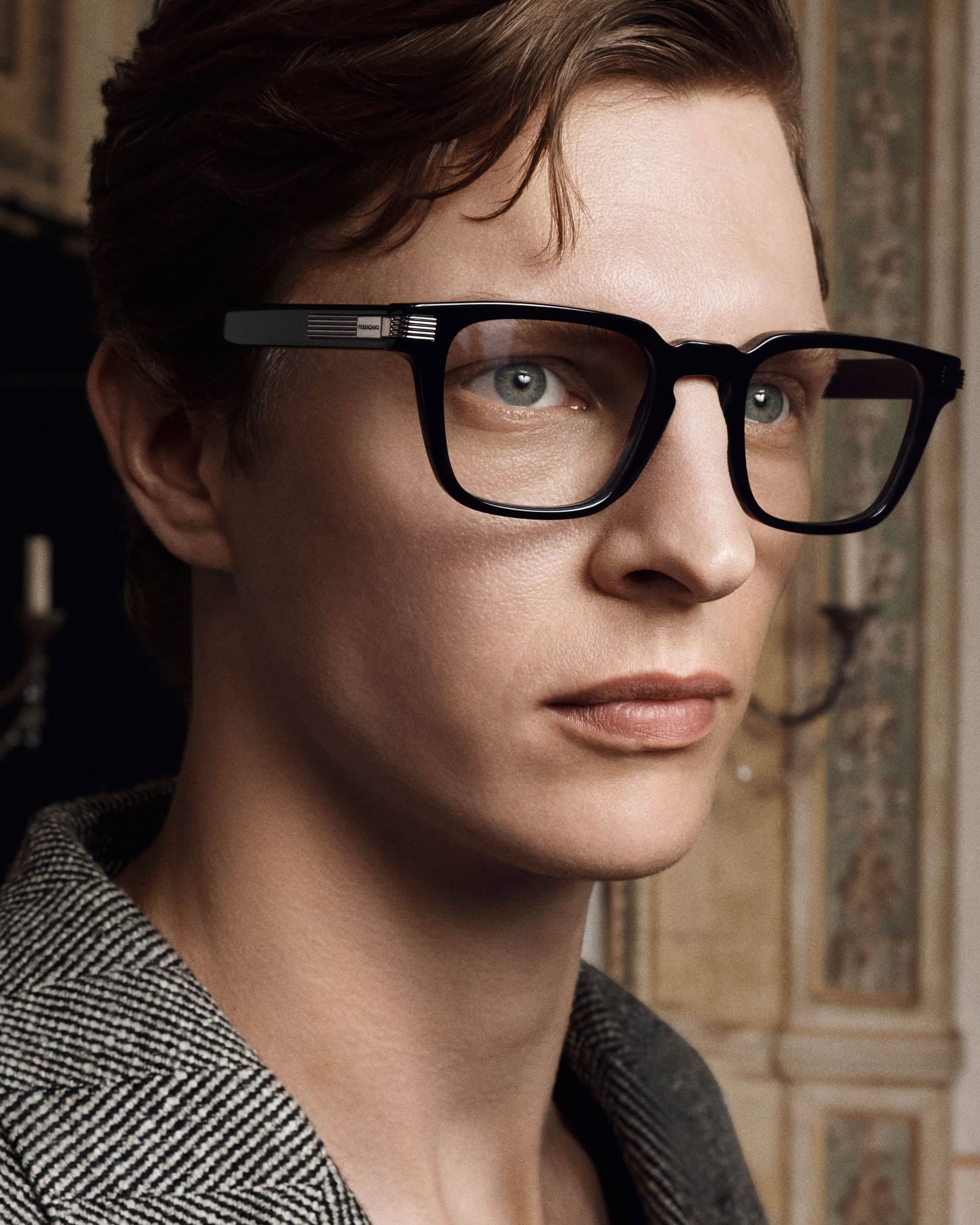 Close-up portrait of a man wearing black-rimmed glasses and a patterned jacket indoors.