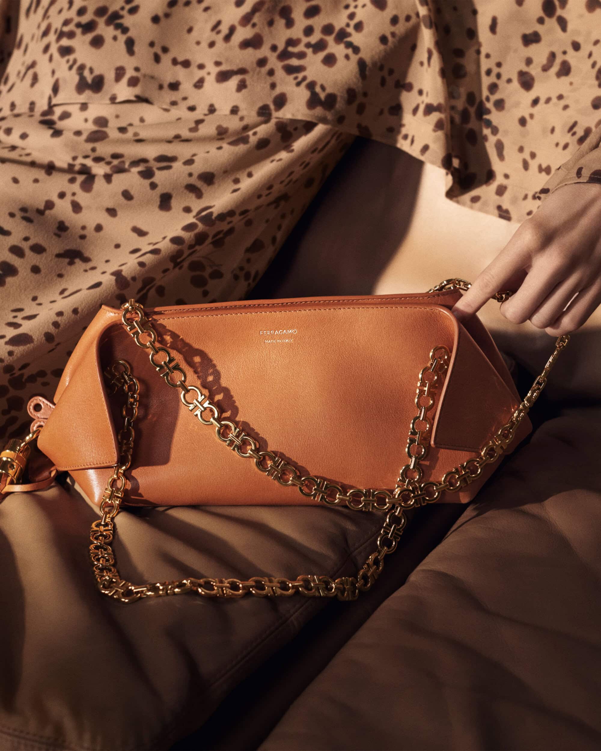 Tan leather shoulder bag with a gold chain strap held by a hand against leopard print fabric.