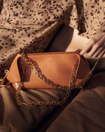 Tan leather shoulder bag with a gold chain strap held by a hand against leopard print fabric.