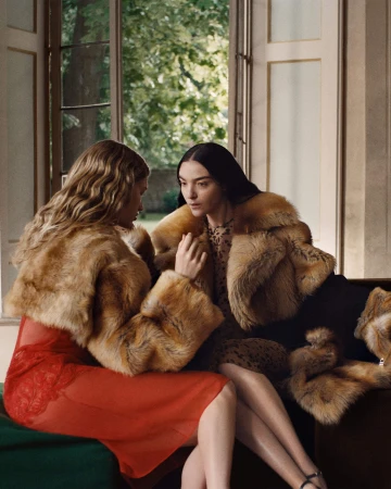 Two women seated indoors wearing fur-collared outerwear and dresses, captured in a campaign photograph by Craig McDean.