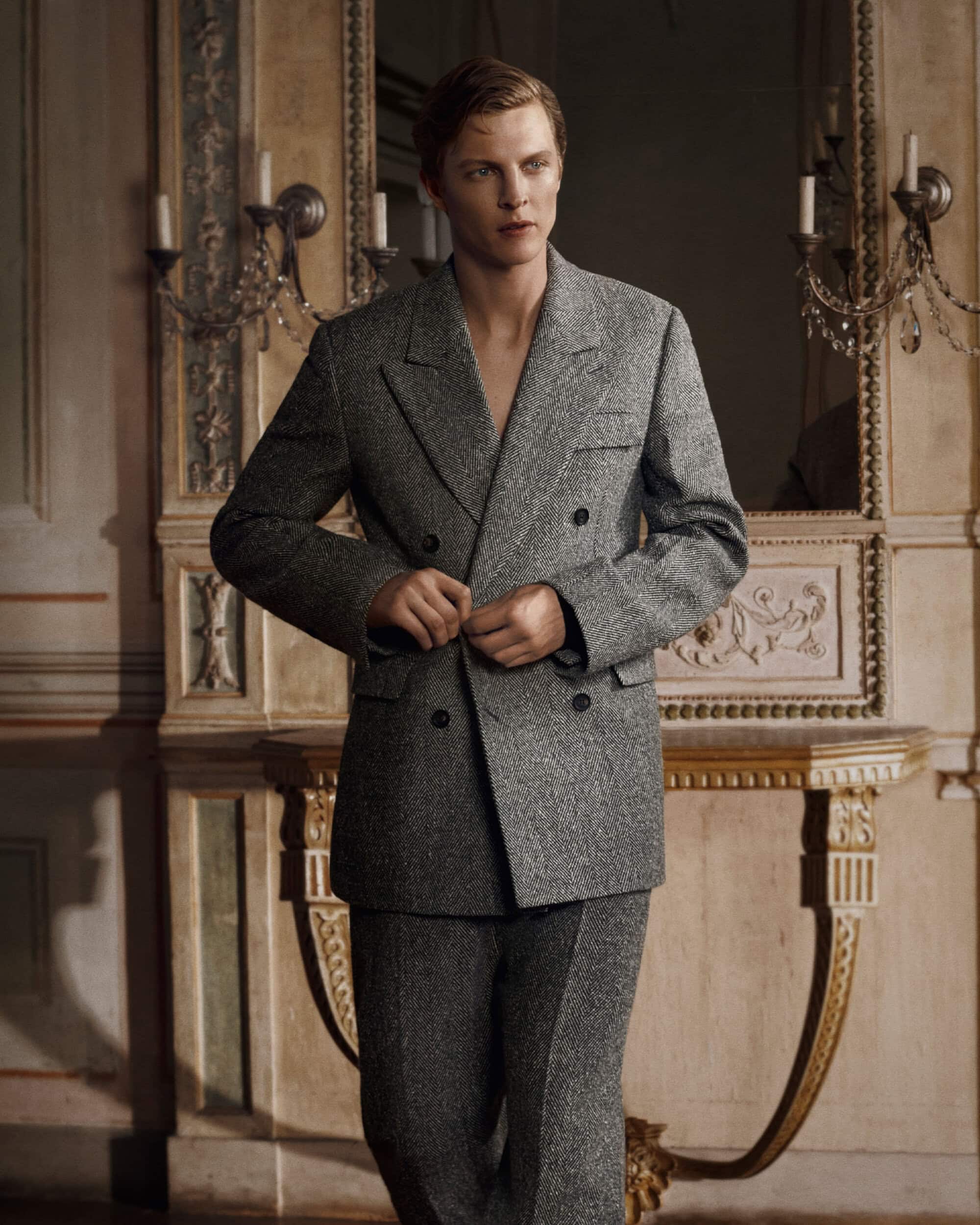 Man wearing a grey tweed double-breasted suit standing in an ornate interior with wall sconces and a mirror.