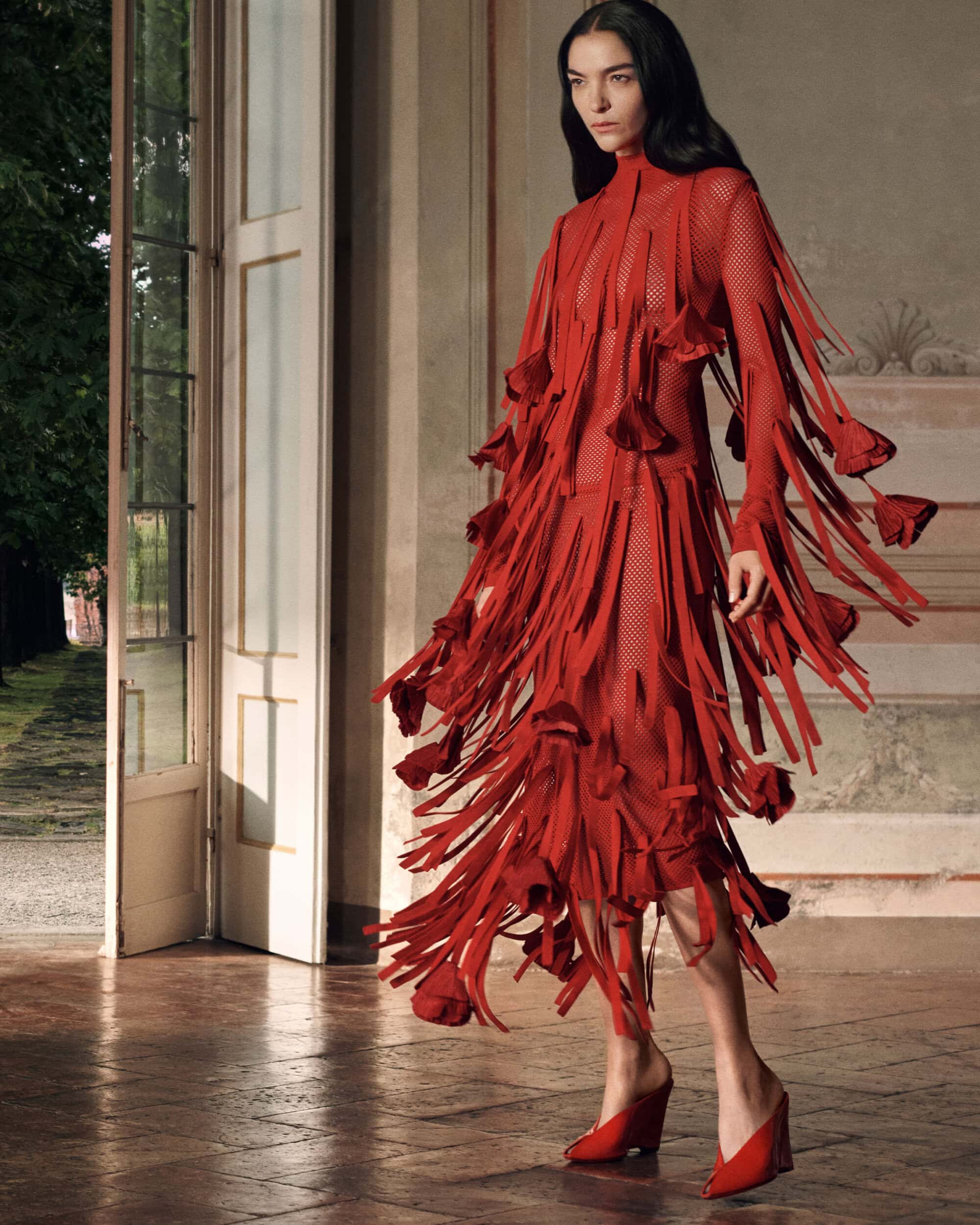 Model in a red fringed dress and red mules standing near an open double door in a classic interior setting.