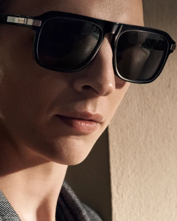 Close-up portrait of a person wearing large black sunglasses and a patterned grey jacket in warm directional light.