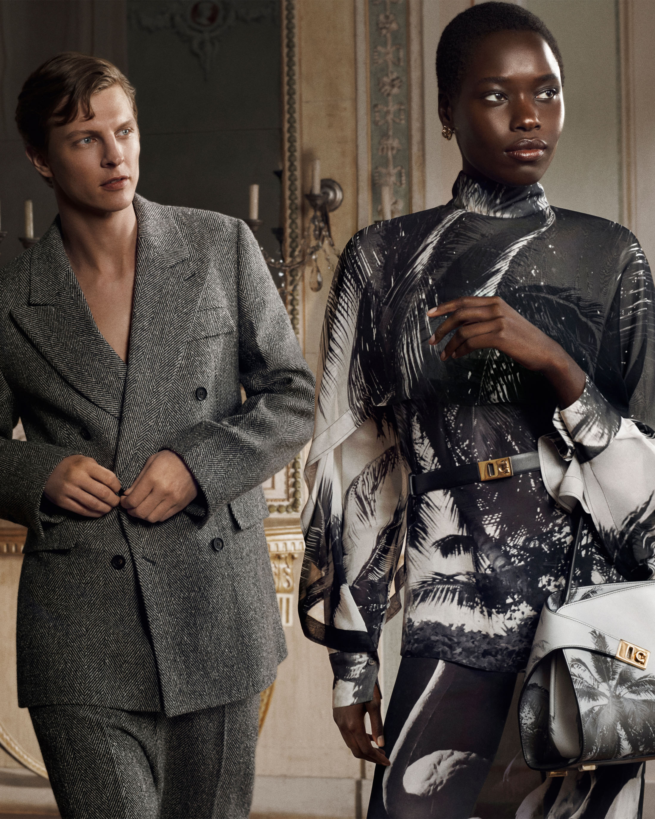 A man in a grey tweed suit standing beside a woman wearing a black and white palm print top and carrying a matching handbag.
