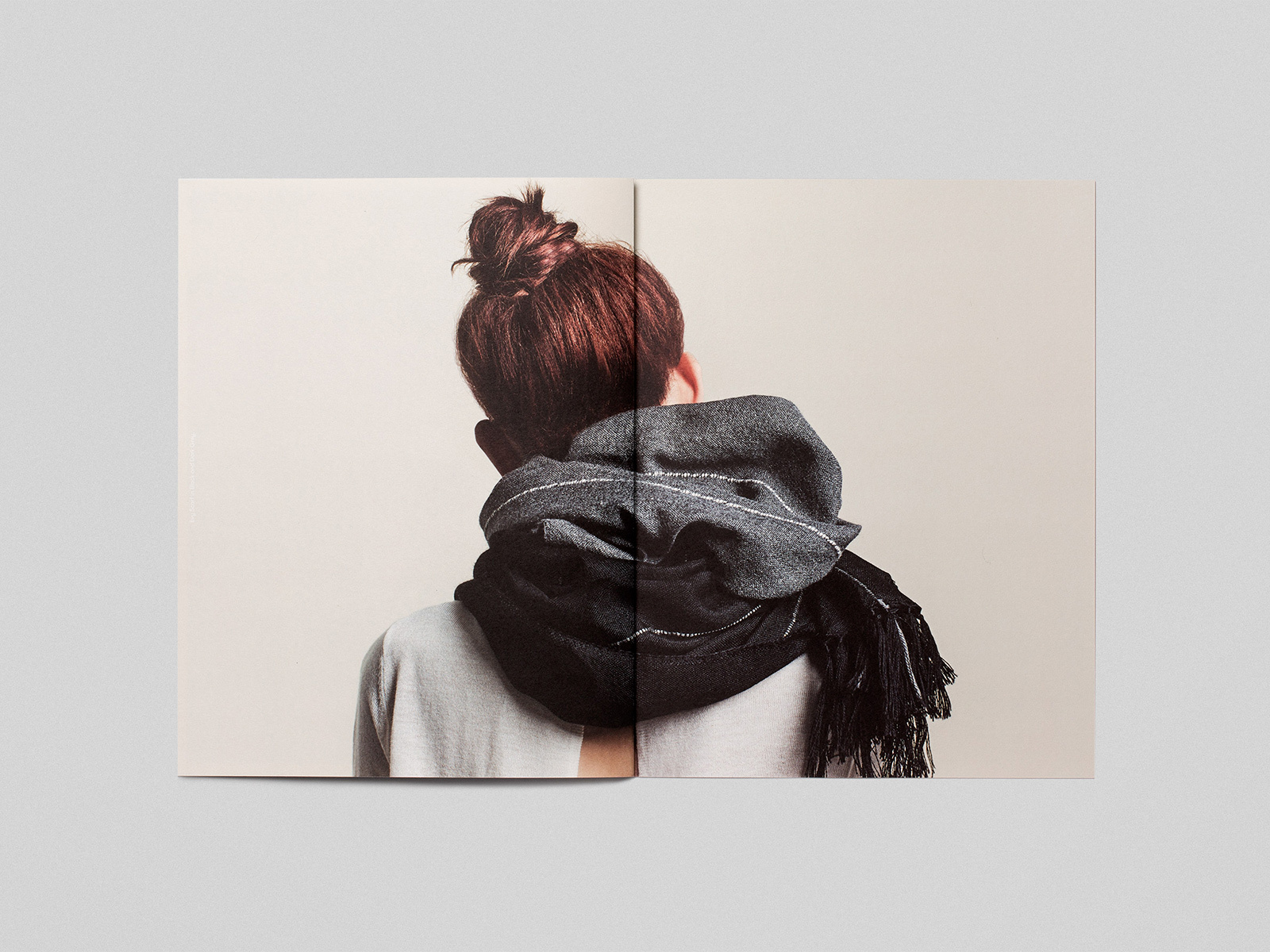 Rear view of a person wearing a dark grey textured scarf and light grey top against a neutral background displayed in an open booklet.