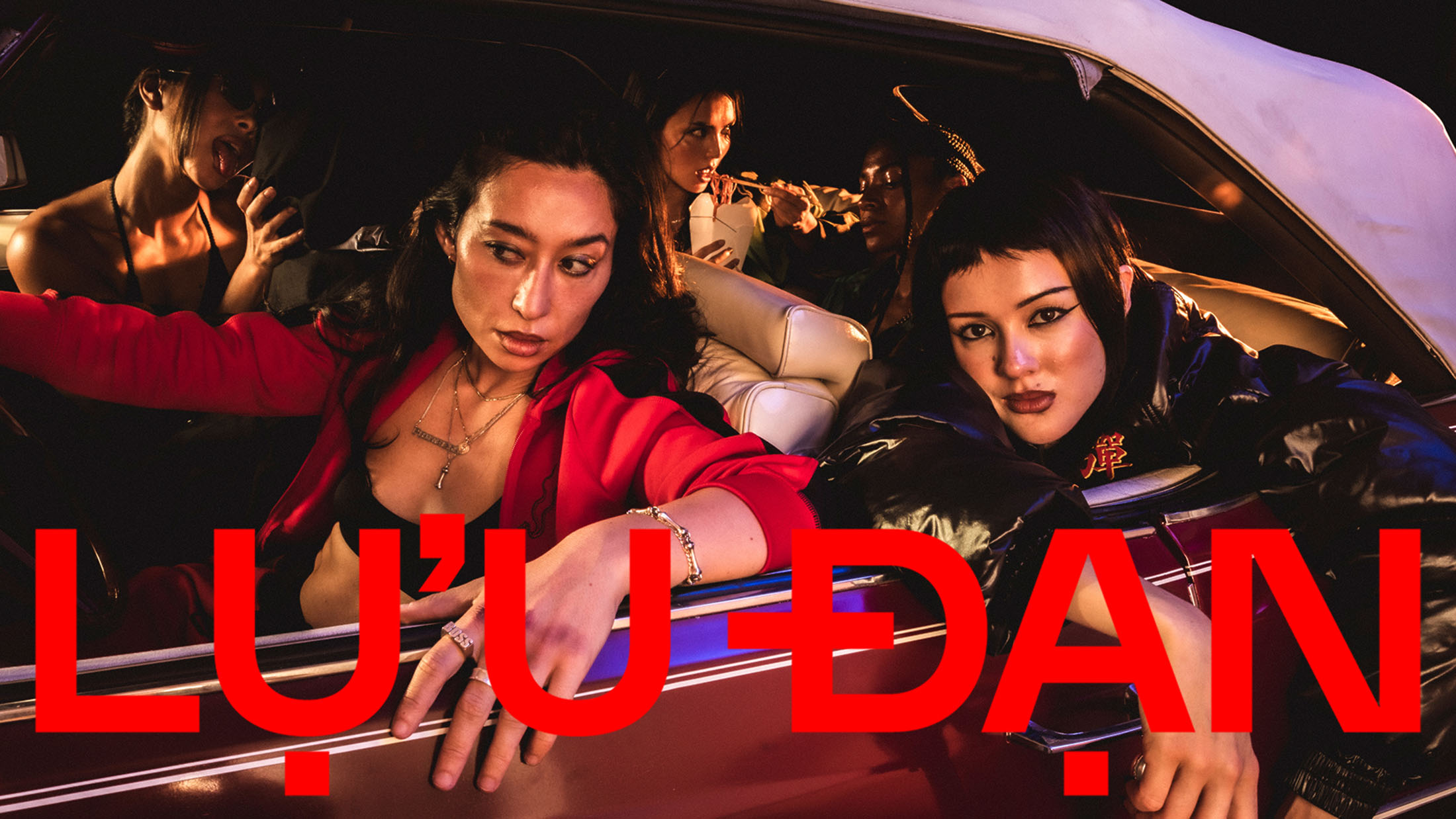 Group of models inside a vintage car under warm lighting for Lu'u Dan Fall 2025 campaign, featuring bold red branding.