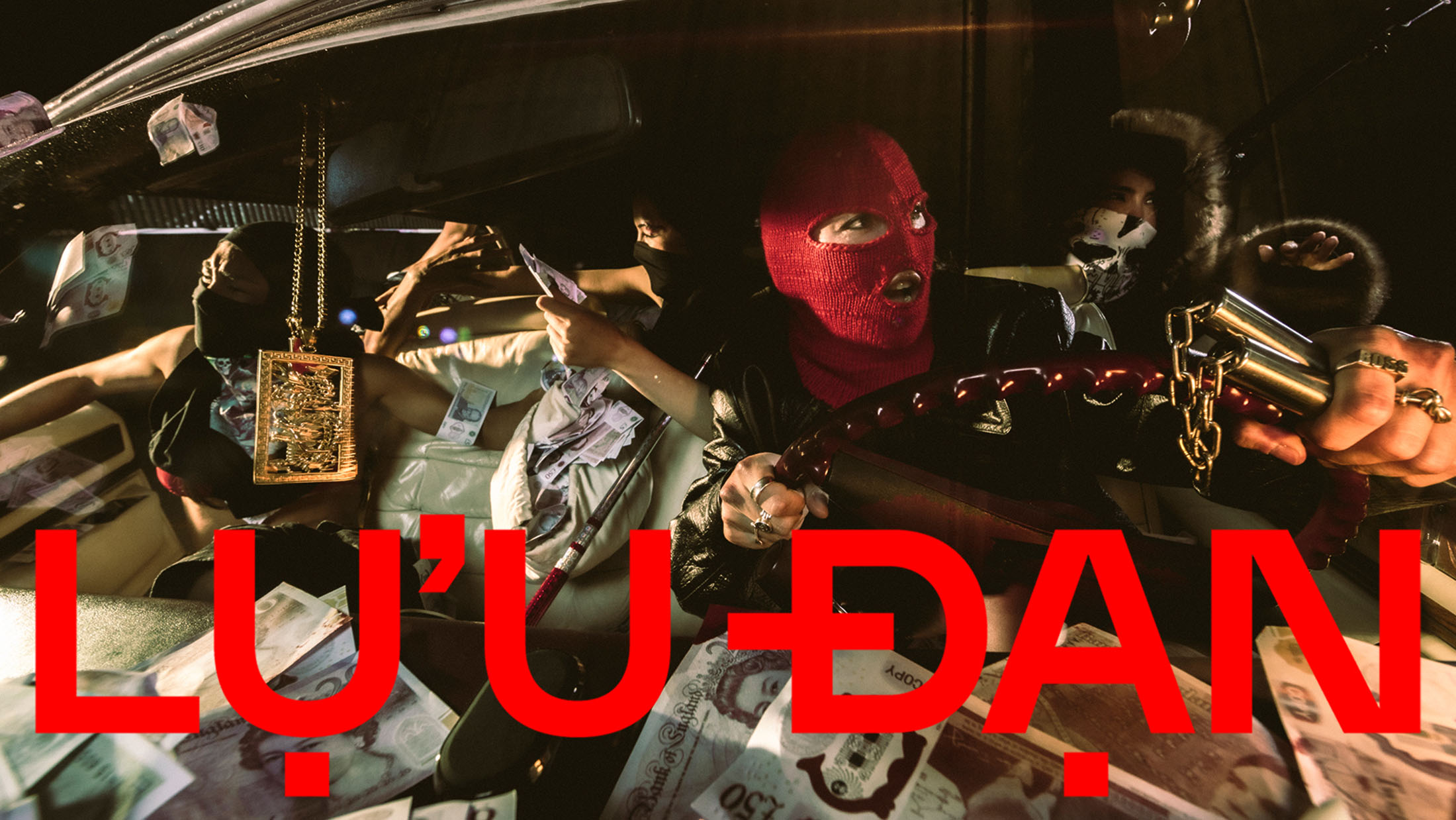 Group of people inside a car, one wearing a red balaclava holding a steering wheel, surrounded by scattered cash and gold jewelry
