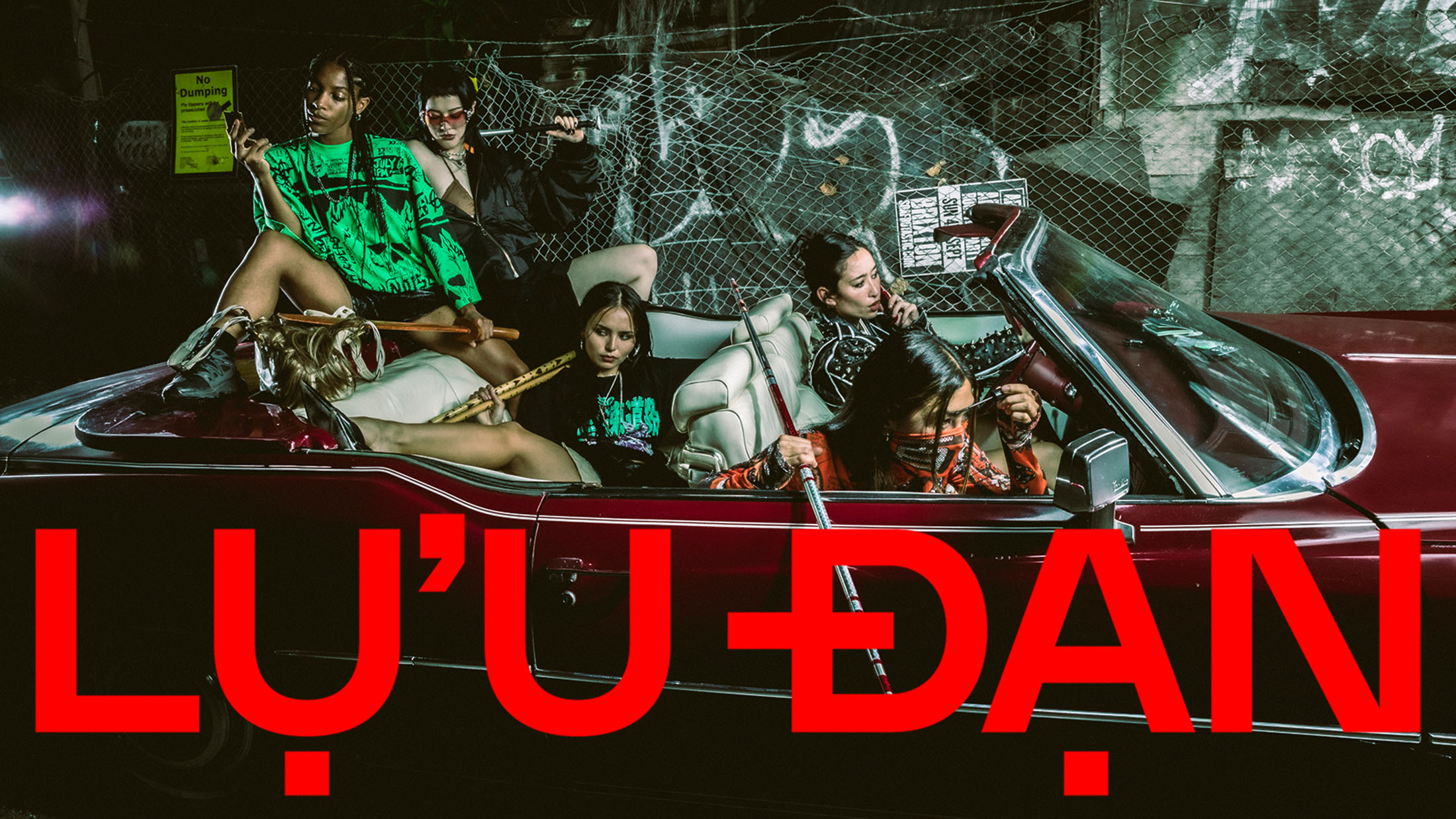 Group of models posing inside a vintage maroon convertible car against a chain-link fence backdrop