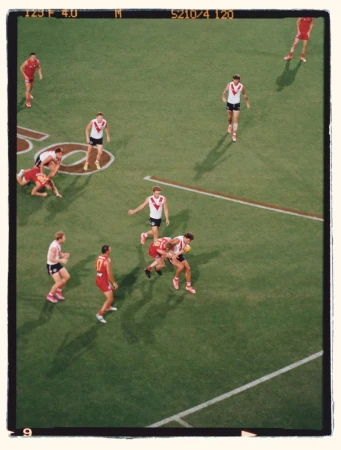 Action shot of an Australian Rules football game on a green field under bright lighting with players in red and white uniforms.