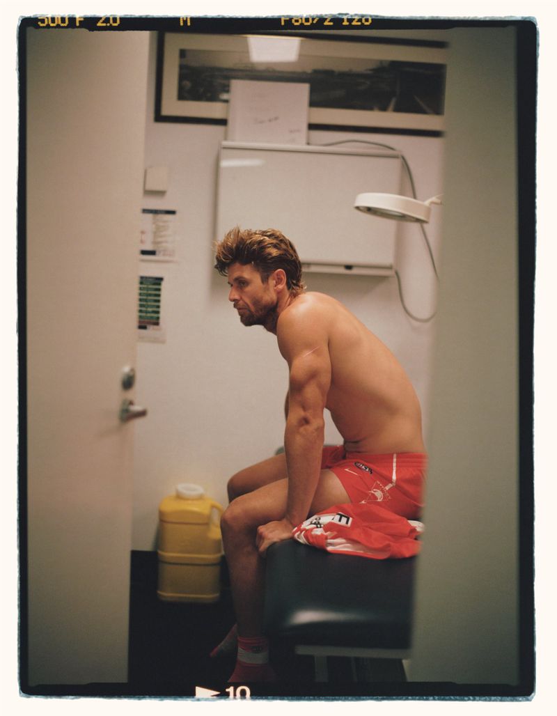 Shirtless male athlete with short hair and muscular build sits on a medical exam table wearing red athletic shorts in an indoor medical environment.