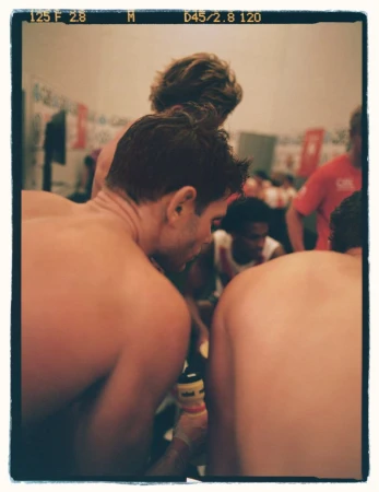 Indoor photo of topless adult men in a locker room, backs and shoulders to camera, with one man's face visible among other athletes.