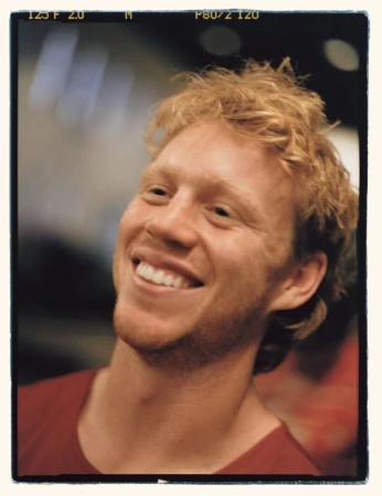Close-up portrait of a smiling adult male with curly blonde hair wearing a red shirt against a dark, blurred background.
