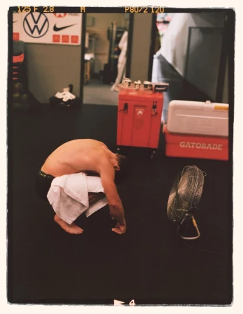Shirtless adult male is bent over with a white towel, in a room with gym equipment, a Gatorade cooler, and sports brand logos.