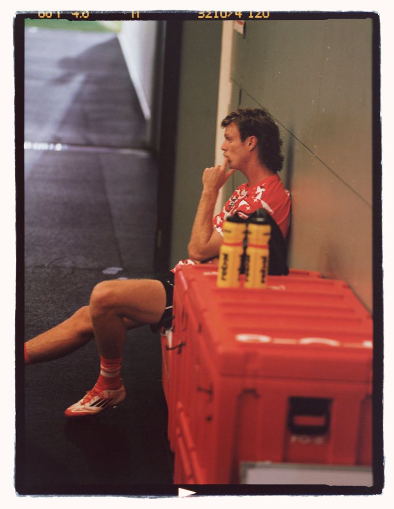 Man in sports gear sits in an indoor training area by a red container, resting and contemplating, post-exercise.